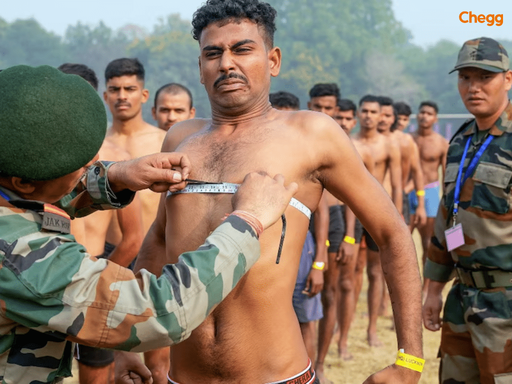 Military personnel in measuring a recruit’s chest during a physical test as others stand in line behind.